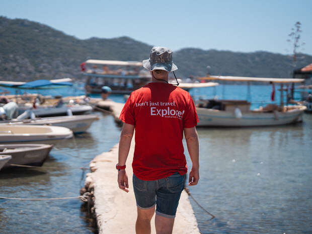 A person in a red shirt with text "Don't just travel. Explore!" walks along a stone pier toward boats on a calm waterfront with hills in the distance under a clear sky.
